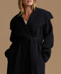 A woman wearing a black wrap coat with a wide collar and self-tie belt, photographed against a solid warm beige background.