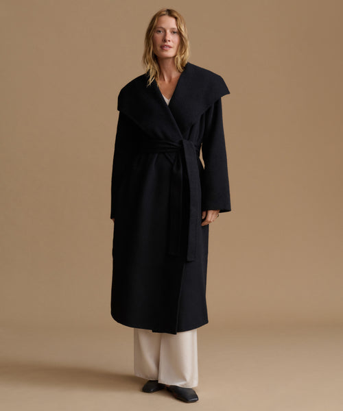 A woman modeling a long black wrap coat with a wide collar and waist tie, paired with cream wide-leg pants and black shoes, against a solid beige studio background.