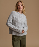 A woman wearing a light gray cable-knit crewneck sweater and olive green pants, standing against a solid warm beige studio background.