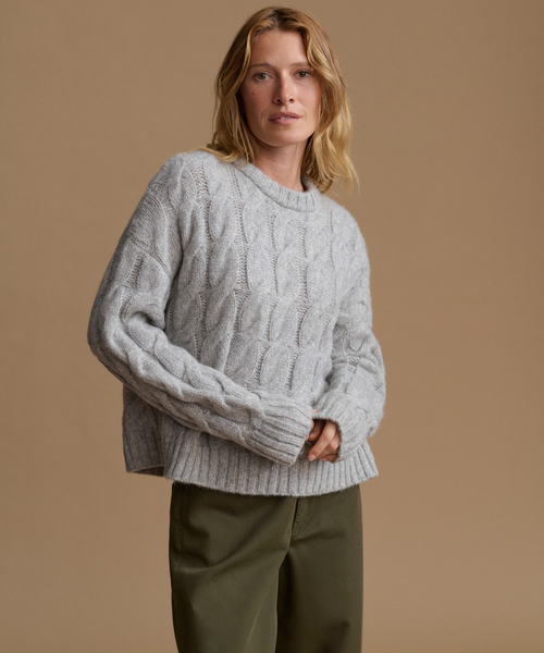 A woman wearing a light gray cable-knit crewneck sweater and olive green pants, standing against a solid warm beige studio background.