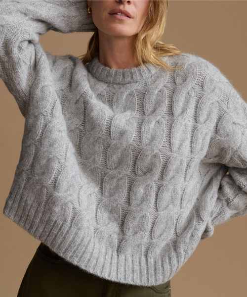 A close-up of a woman wearing a light gray cable-knit sweater with a crew neck, paired with olive green pants, against a solid warm beige background.