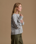 A woman in profile wearing a light gray chunky cable-knit sweater and olive green pants, posed against a solid warm beige studio background.