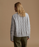 A woman shown from the back wearing a light gray cable-knit sweater and olive green pants, set against a solid warm beige studio background.
