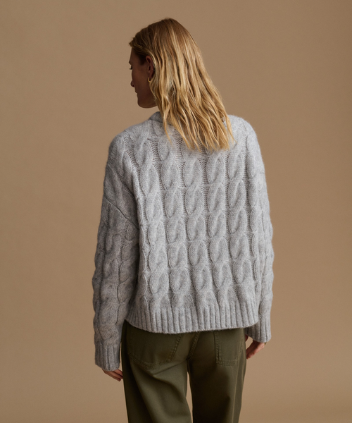 A woman shown from the back wearing a light gray cable-knit sweater and olive green pants, set against a solid warm beige studio background.
