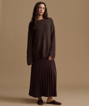 A woman stands against a plain warm beige studio background wearing an oversized dark brown knit sweater, a matching dark brown pleated maxi skirt, and dark brown slip-on shoes.