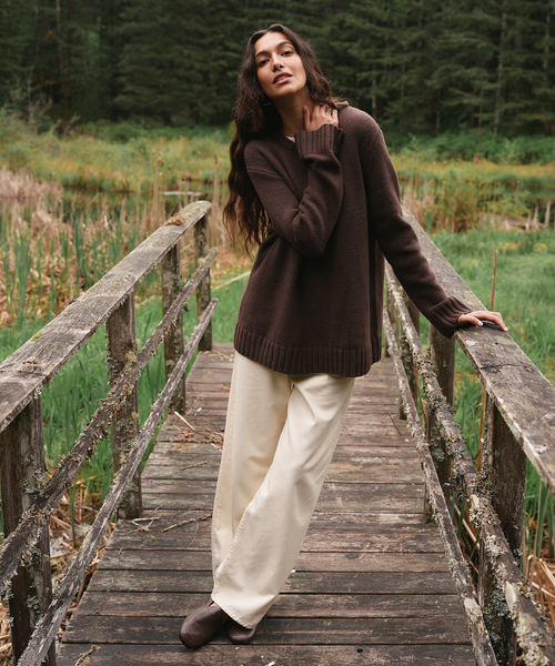 A woman stands on a rustic wooden bridge in a grassy forest clearing, wearing an oversized dark brown knit sweater, cream wide-leg pants, and dark brown boots.