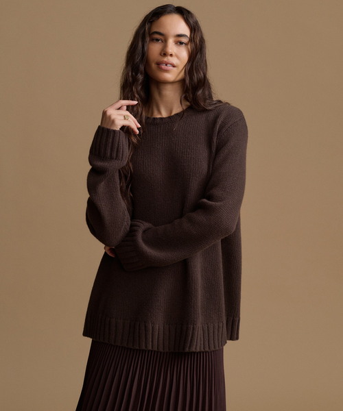 A woman modeling a dark brown ribbed knit sweater with a crew neck and long sleeves, paired with a matching pleated skirt, against a solid warm brown studio background.