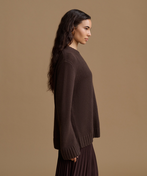 A woman in side profile wearing a dark brown knit sweater and a pleated dark skirt, set against a solid warm tan studio background.