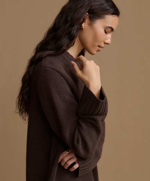 A woman in side profile wearing a dark brown textured knit sweater against a solid warm beige background.