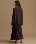 A woman shown from the back wearing a dark brown knit sweater and a matching dark brown pleated maxi skirt, standing against a solid beige studio background.