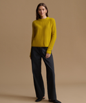 A woman modeling a ribbed mustard yellow crewneck sweater paired with dark charcoal wide-leg trousers, standing against a solid warm beige studio background.