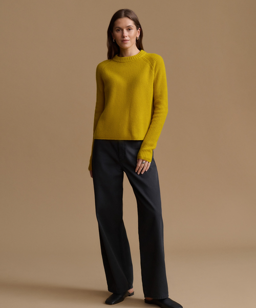 A woman modeling a ribbed mustard yellow crewneck sweater paired with dark charcoal wide-leg trousers, standing against a solid warm beige studio background.