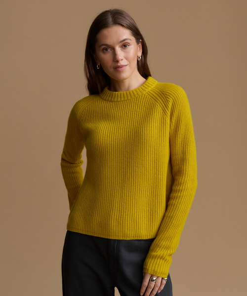 A woman wearing a ribbed knit mustard yellow sweater and black pants, standing against a solid warm beige studio background.