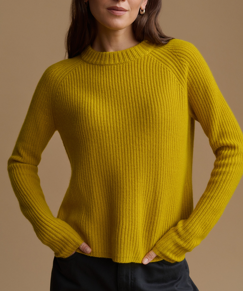A woman wearing a ribbed knit crewneck sweater in a rich mustard yellow color, paired with dark trousers, against a solid warm beige background.