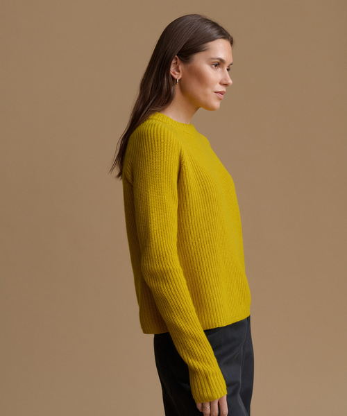 A woman in side profile wearing a ribbed knit mustard yellow sweater and dark trousers, set against a solid warm tan studio background.