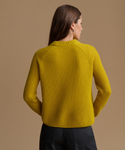 A woman shown from the back wearing a ribbed knit sweater in a rich mustard yellow color, paired with dark trousers, against a solid warm beige studio background.