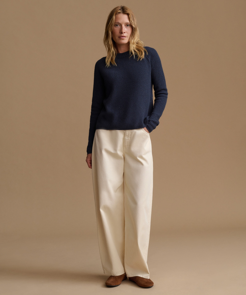 A woman modeling a dark navy ribbed knit sweater with long sleeves paired with cream wide-leg pants, against a solid warm beige studio background.