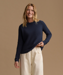 A woman modeling a dark navy ribbed knit sweater with long sleeves paired with cream wide-leg pants, against a solid warm beige studio background.