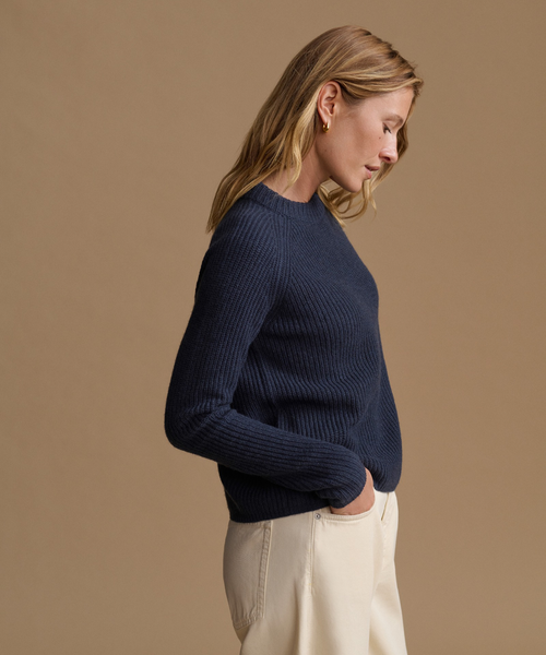 A woman in side profile wearing a navy blue ribbed knit sweater and cream-colored trousers, set against a solid warm beige studio background.