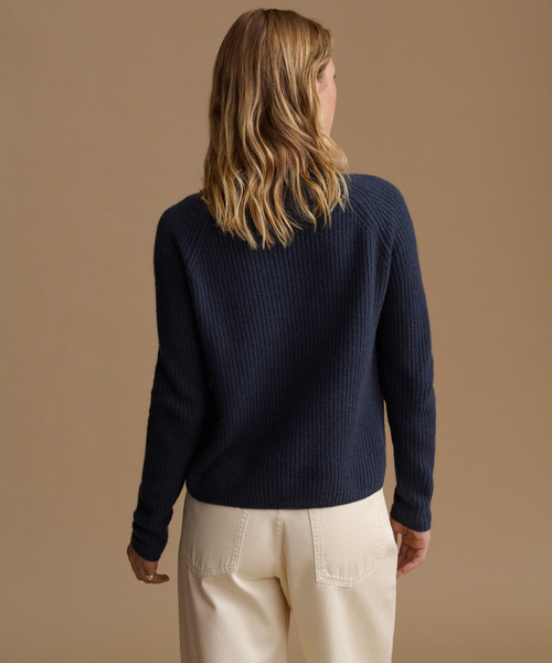 A woman modeling a dark navy ribbed knit sweater with long sleeves paired with cream wide-leg pants, against a solid warm beige studio background.