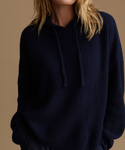 A close-up of a person wearing an oversized navy blue ribbed knit hoodie with matching drawstrings, against a warm beige studio background.