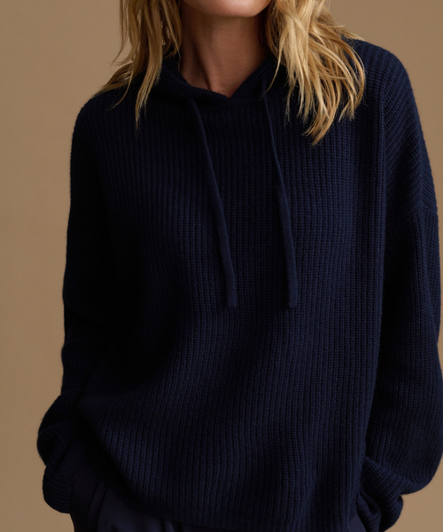 A close-up of a person wearing an oversized navy blue ribbed knit hoodie with matching drawstrings, against a warm beige studio background.