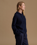 A woman in profile wearing a dark navy ribbed knit hoodie and matching navy pants, standing against a solid warm beige studio background.