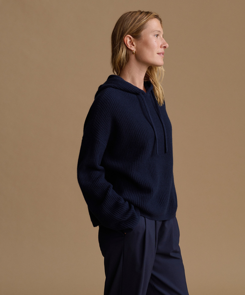 A woman in profile wearing a dark navy ribbed knit hoodie and matching navy pants, standing against a solid warm beige studio background.
