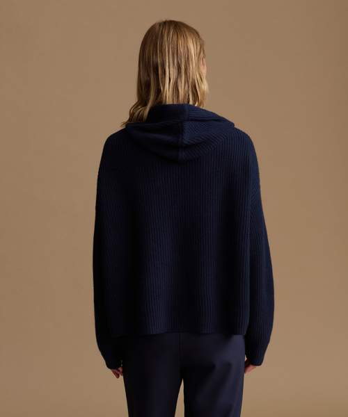 A model shown from the back wearing a navy blue ribbed knit hooded sweater and matching navy trousers, set against a solid warm beige studio background.