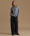 A model stands against a solid tan studio background wearing a textured gray cropped hoodie with drawstrings and black wide-leg trousers.