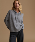 A woman wearing a ribbed gray knit hoodie sweater and dark charcoal pants, standing against a solid warm beige studio background.