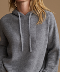 A close-up of a woman wearing a gray ribbed knit hooded sweater with matching drawstrings, set against a solid warm beige background.