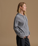 A woman in profile wearing a ribbed gray knit hoodie and dark pants, standing against a plain warm beige studio background.