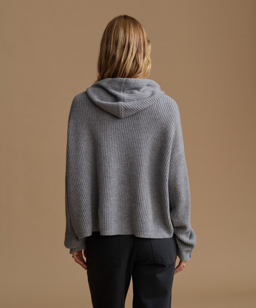 A woman viewed from behind wearing a gray ribbed knit cropped hoodie with long sleeves and black pants, standing against a solid warm tan background.