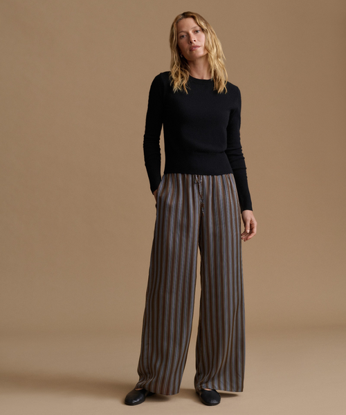 A woman stands against a plain warm beige studio background wearing a fitted black long-sleeve top and wide-leg striped pants in brown and gray tones, with black shoes.