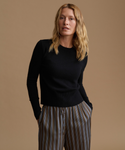 A woman modeling a black long-sleeve knit sweater with striped trousers, standing against a solid warm beige studio background.