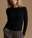 A woman stands against a plain warm beige studio background wearing a fitted black long-sleeve top and wide-leg striped pants in brown and gray tones, with black shoes.