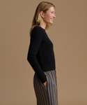 A woman in side profile wearing a black long-sleeve knit top and striped trousers against a solid tan studio background.