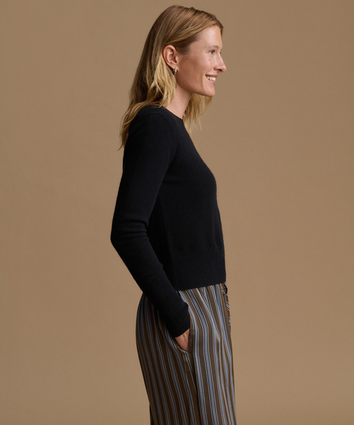 A woman in side profile wearing a black long-sleeve knit top and striped trousers against a solid tan studio background.