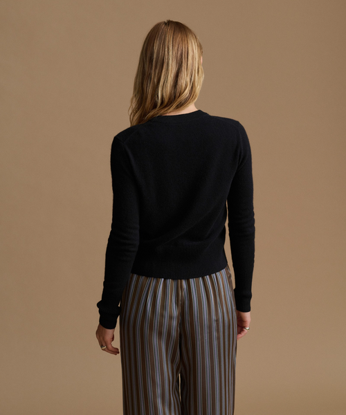 A woman shown from the back wearing a black long-sleeve knit top and striped trousers, standing against a solid warm beige studio background.