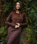 A woman wearing a long-sleeve brown knit dress stands against a mossy tree trunk in a forest setting.