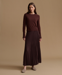 A woman stands against a plain warm beige studio background wearing a dark brown long-sleeve knit top and a matching dark brown pleated maxi skirt, with taupe slip-on shoes.