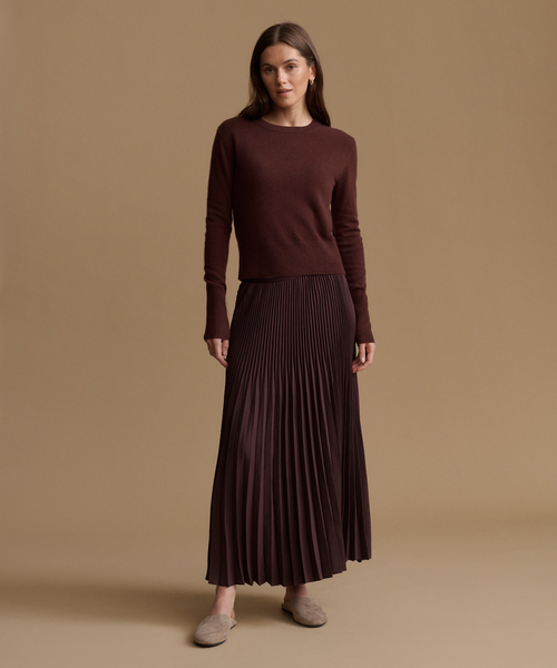 A woman stands against a plain warm beige studio background wearing a dark brown long-sleeve knit top and a matching dark brown pleated maxi skirt, with taupe slip-on shoes.