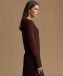 A woman in side profile wearing a dark brown ribbed knit sweater and a dark pleated skirt, set against a solid warm beige studio background.