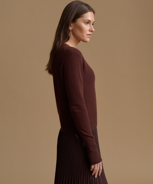 A woman in side profile wearing a dark brown ribbed knit sweater and a dark pleated skirt, set against a solid warm beige studio background.