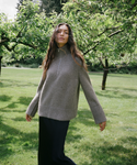 A woman standing in a grassy park under leafy trees, wearing a loose taupe-gray ribbed knit mock-neck sweater and dark wide-leg pants.