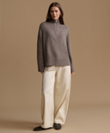 A woman stands against a plain warm beige studio background wearing a taupe-gray half-zip mock-neck knit sweater, cream wide-leg trousers, and dark slip-on shoes.