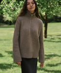 A woman wearing a taupe brown ribbed knit mock neck sweater with a short front zipper, standing in a grassy park with trees in the background.