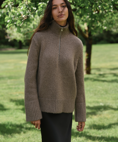 A woman wearing a taupe brown ribbed knit mock neck sweater with a short front zipper, standing in a grassy park with trees in the background.