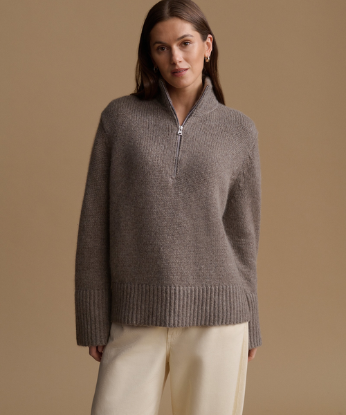 A woman stands against a plain warm beige studio background wearing a taupe-gray half-zip mock-neck knit sweater, cream wide-leg trousers, and dark slip-on shoes.